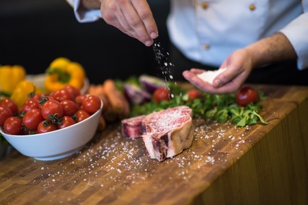 Master Chef hands putting salt on juicy slice of raw steak with vegetables around on a wooden tableの写真素材
