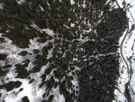 top view aerial drone flying above curvy road wind mountains and forest covered with snow on bad weatherの写真素材