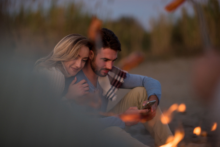 Group of young friends sitting by the fire at autumn beach, grilling sausages and drinking beer, talking and having funの写真素材