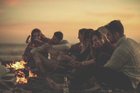 Group of young friends sitting by the fire at autumn beach, grilling sausages and drinking beer,...