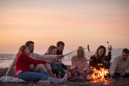 Group of young friends sitting by the fire at autumn beach, grilling sausages and drinking beer, talking and having funの写真素材