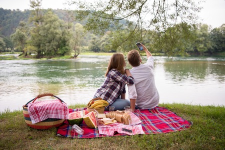 Couple in love taking a selfie by mobile phone while enjoying picnic time drink and food in beautiful nature on the river bankの写真素材