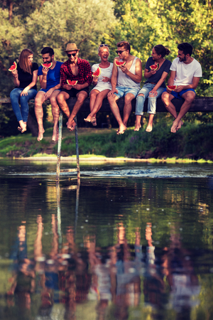 group of young friends enjoying watermelon while sitting on the wooden bridge over the river in beautiful natureの写真素材