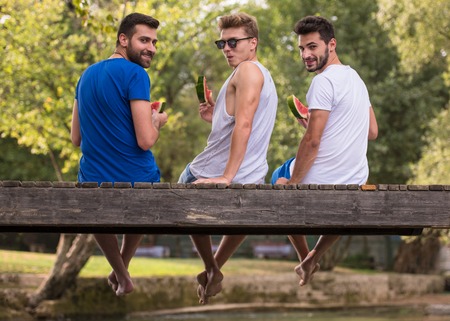 group of young men enjoying watermelon while sitting on the wooden bridge over the river in beautiful natureの写真素材