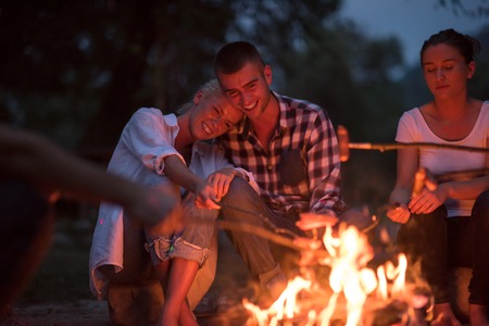 a group of happy young friends relaxing and enjoying  summer evening around campfire on the river bankの写真素材