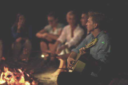 a group of happy young friends relaxing and enjoying  summer evening around campfire on the river...