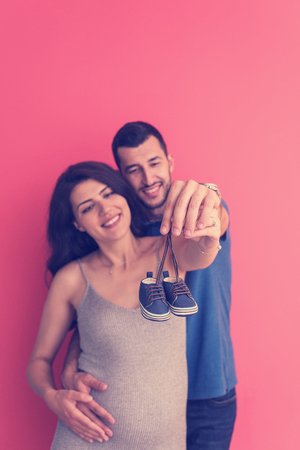 young  pregnant couple holding newborn baby shoes isolated on red background,family and parenthood conceptの写真素材