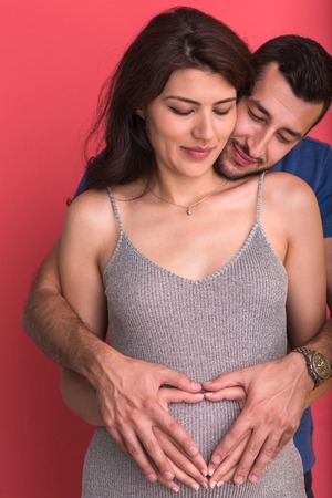 Happy young couple showing heart with their hands on belly of pregnant wife isolated on red backgroundの写真素材