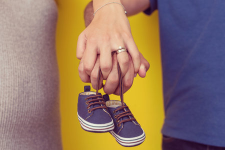 young  pregnant couple holding newborn baby shoes isolated on yellow background,family and...