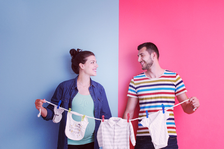Beautiful pregnant woman and her husband expecting baby holding baby bodysuits and smiling over colorful backgroundの写真素材