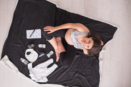 A pregnant woman at home on the floor checking list of baby clothes preparing for going to maternity...