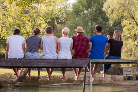 A group of young friends enjoying watermelon while sitting on the wooden bridge over the river in beautiful nature rear viewの写真素材