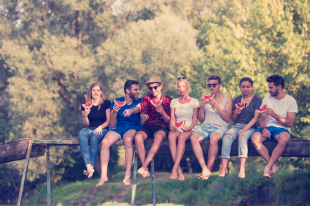 A group of young friends enjoying watermelon while sitting on the wooden bridge over the river in beautiful natureの写真素材