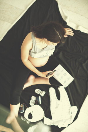 A pregnant woman at home on the floor checking list of baby clothes preparing for going to maternity...