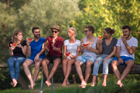 A group of young friends enjoying watermelon while sitting on the wooden bridge over the river in beautiful natureの写真素材