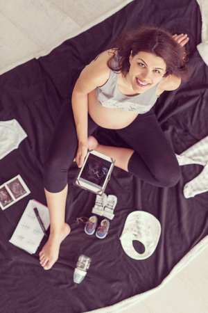 A pregnant woman at home on the floor checking list of baby clothes preparing for going to maternity hospital top view.の写真素材