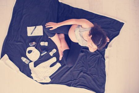 A pregnant woman at home on the floor checking list of baby clothes preparing for going to maternity hospital top view.の写真素材