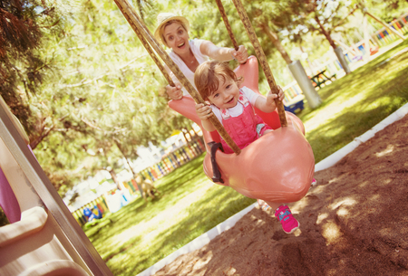 young mother and her little daughter smiling together while swinging at the playground in the parkの写真素材