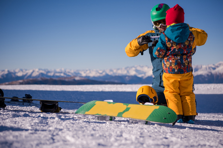 Young happy father preparing his little son for the first time on a snowboard during sunny winter day at beautiful  ski resortの写真素材