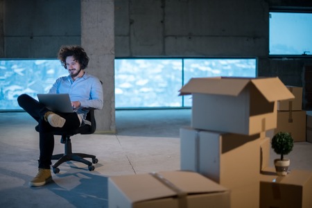 Young male architect on construction site checking documents and business workflow using laptop computer with cardboard boxes around him in new startup officeの写真素材