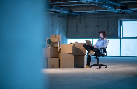 Young male architect on construction site checking documents and business workflow using laptop computer with cardboard boxes around him in new startup officeの写真素材