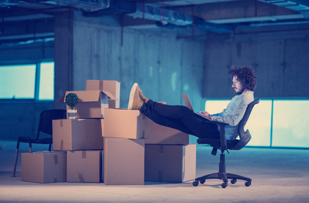 Young male architect on construction site checking documents and business workflow using laptop computer with cardboard boxes around him in new startup officeの写真素材