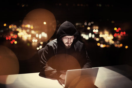 Young talented hacker using laptop computer while working in dark office with big city lights
