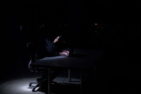 Young talented hacker using laptop computer while working in dark office with big city lightsの写真素材