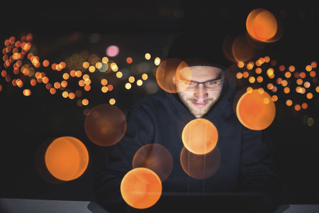 Young talented hacker using laptop computer while working in dark office with big city lights