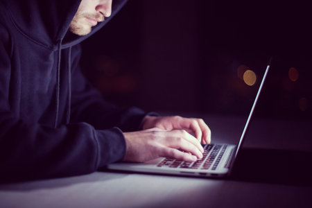 Young talented hacker using laptop computer while working in dark office with big city lights