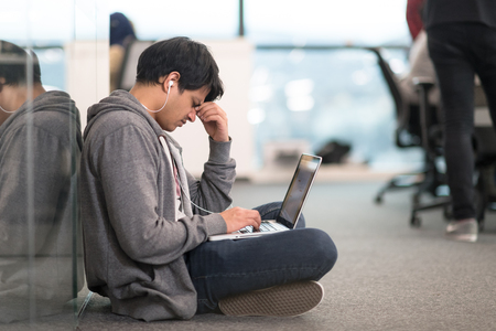 Young Indian software developer man using laptop computer writing programming code while sitting on the floor at modern creative startup officeの写真素材