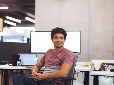Portrait of young indian successful male software developer sitting on desk at modern startup office