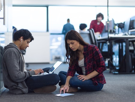 Young software developers couple using laptop and tablet computer writing programming code while sitting on the floor at modern creative startup officeの写真素材