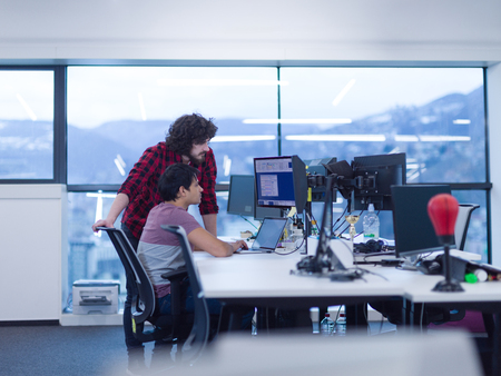 Two young male software developers using laptop and desktop computer while writing programming code at modern creative startup officeの写真素材