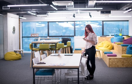 Young redhead businesswoman reading a text message on her mobile phone while having a break in the modern startup officeの写真素材
