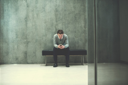 Young businessman using smart phone while sitting on the bench at office lobby during a breakの写真素材