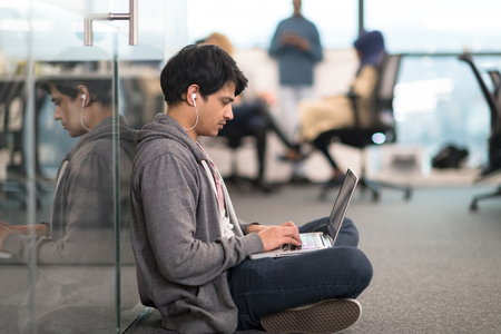 young indian software developer man using laptop computer writing programming code while sitting on the floor at modern creative startup officeの写真素材