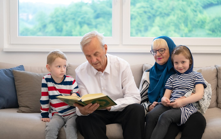 modern muslim family grandparents with grandchildren reading Quran and praying together on the sofa before iftar dinner during a ramadan feast at homeの写真素材