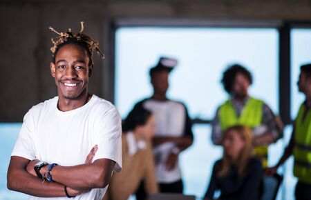 portrait of young casual african american businessman standing with crossed arms and multiethnic business team in the background on construction site  during moving in at new startup officeの写真素材