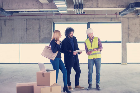 architect using tablet computer while showing house design plans to a young couple at construction...