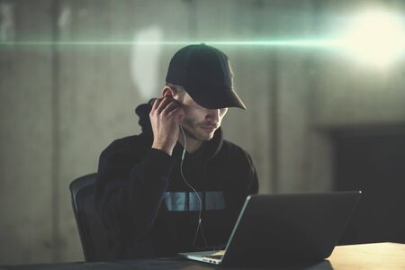 Young talented hacker using laptop computer while working in dark office with concrete wall and light flare in the backgroundの写真素材