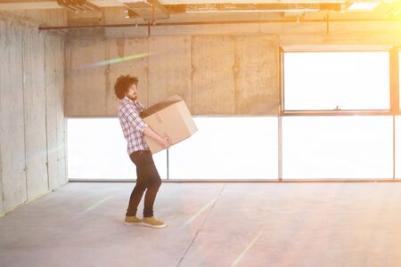 happy young casual business man carrying cardboard box with sunlight through the windows during moving in at new unfinished startup office buildingの写真素材