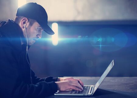 Young talented hacker using laptop computer while working in dark office with concrete wall and light flare in the backgroundの写真素材