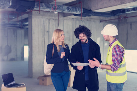 architect using tablet computer while showing house design plans to a young couple at construction...