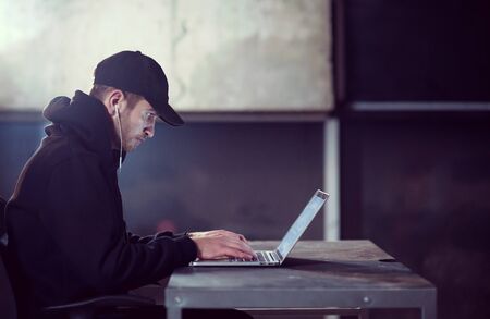 Young talented hacker using laptop computer while working in dark office with concrete wall in the backgroundの写真素材