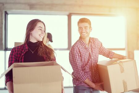 happy young casual business team carrying cardboard boxes with sunlight through the windows during moving in at new unfinished startup office buildingの写真素材