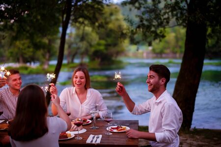 group of happy friends celebrating holiday vacation using sprinklers and drinking red wine while having picnic french dinner party outdoor near the river on beautiful summer evening in natureの写真素材