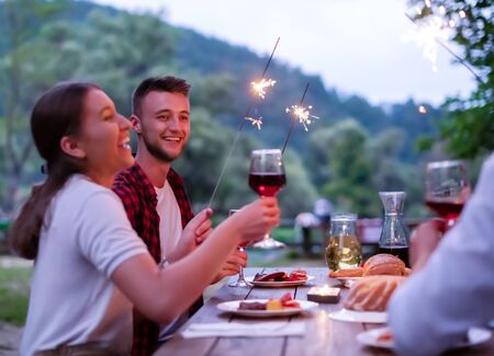 group of happy friends celebrating holiday vacation using sprinklers and drinking red wine while having picnic french dinner party outdoor near the river on beautiful summer evening in natureの写真素材