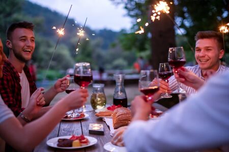 group of happy friends celebrating holiday vacation using sprinklers and drinking red wine while having picnic french dinner party outdoor near the river on beautiful summer evening in natureの写真素材