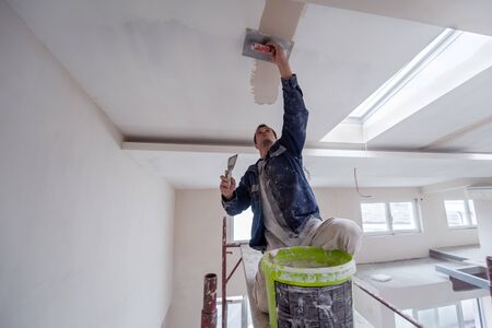 young professional construction worker using scaffold while plastering on gypsum ceiling inside the new big modern two levels apartmentの写真素材